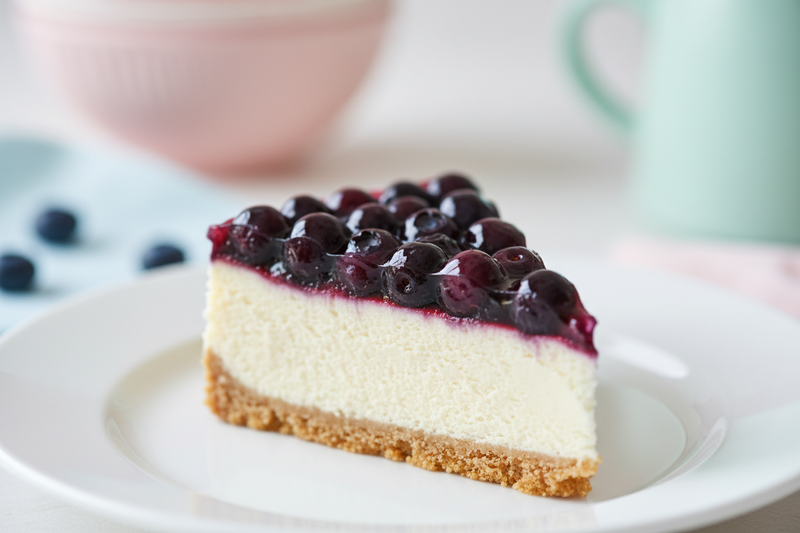 A close-up slice of blueberry cheesecake topped with fresh plump blueberries and a golden graham cracker crust, served on a white ceramic plate